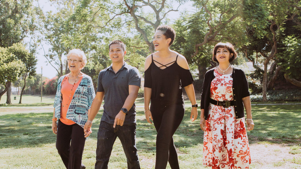 Four people of diverse ages and ethnicities walking together in a sunny park, smiling and holding hands.