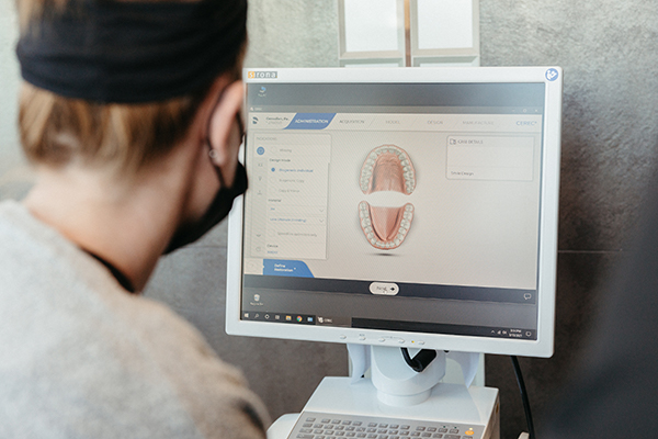 Person wearing a black beanie views a dental software program displaying 3d teeth models on a computer monitor in an office setting.