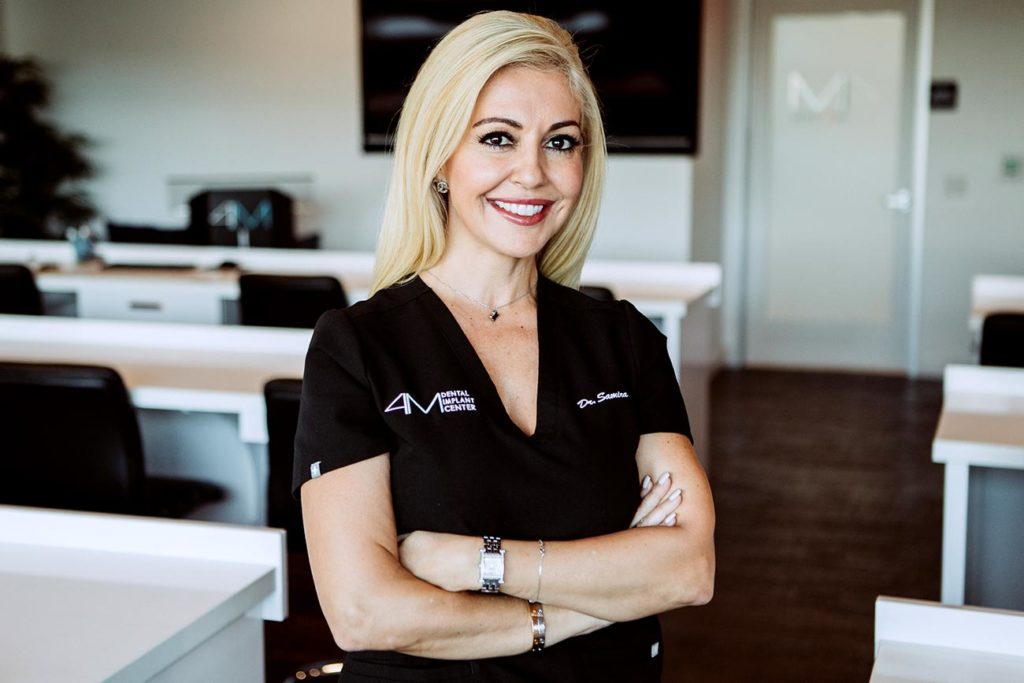 A professional woman with blonde hair, showcasing her cosmetic dentistry smile, in an office environment, wearing a black shirt with a name tag, arms crossed.