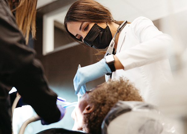 A dentist wearing a mask and gloves examines a patient's teeth for whole mouth dental implants while an assistant helps during the procedure.