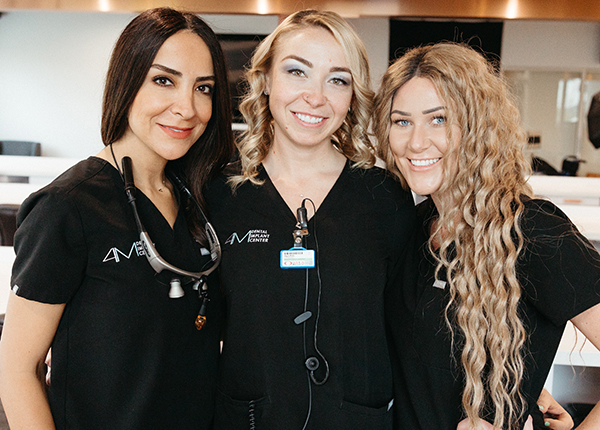 Three female dental professionals specializing in whole mouth dental implants, smiling in an office setting.