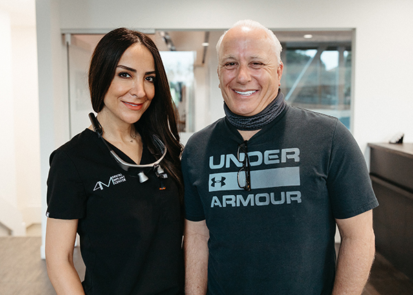 A man and a woman with whole mouth dental implants smiling at the camera inside an office, the woman wearing a black work uniform and the man in a grey under armour t-shirt.