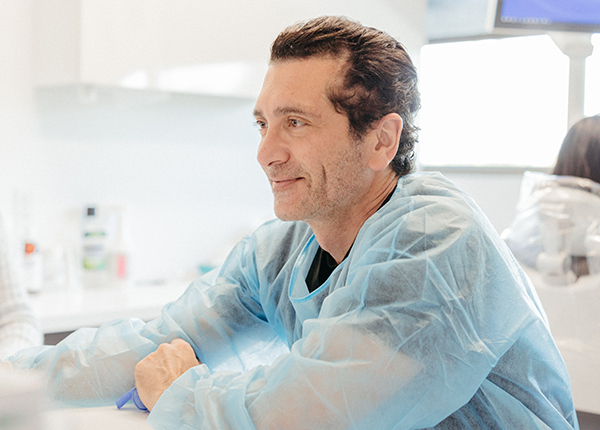 A male medical professional specializing in whole mouth dental implants, wearing a blue scrub suit, sits smiling in a bright clinical laboratory setting.