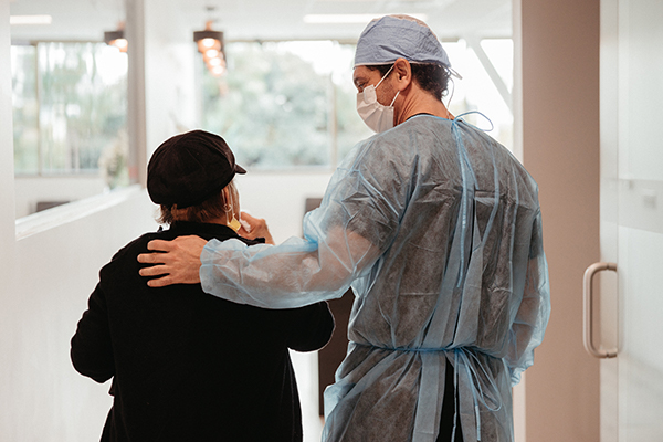 A healthcare worker in scrubs and a mask comforting an elderly patient with whole mouth dental implants in a hospital corridor.