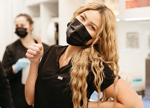 Blonde woman in a black mask and uniform giving a thumbs-up in a dental clinic specializing in whole mouth dental implants, with another masked person in the background.