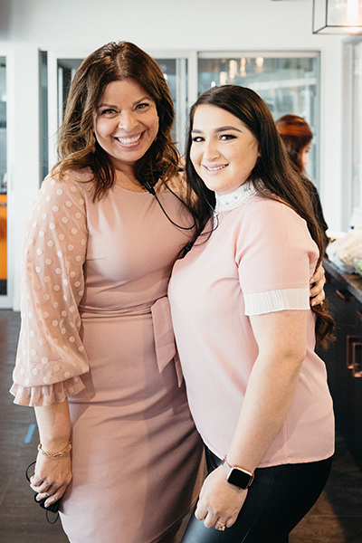 Two women smiling with whole mouth dental implants and embracing inside a modernly styled venue, both dressed in pink outfits.