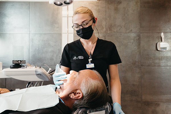 A female dermatologist in a mask examines an elderly male patient's face, focusing particularly on the area around his whole mouth dental implants, in a clinical setting.