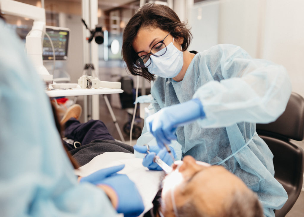 Dentist in blue scrubs and mask examining a patient's teeth in a dental clinic, using tools, with dental equipment in the background.