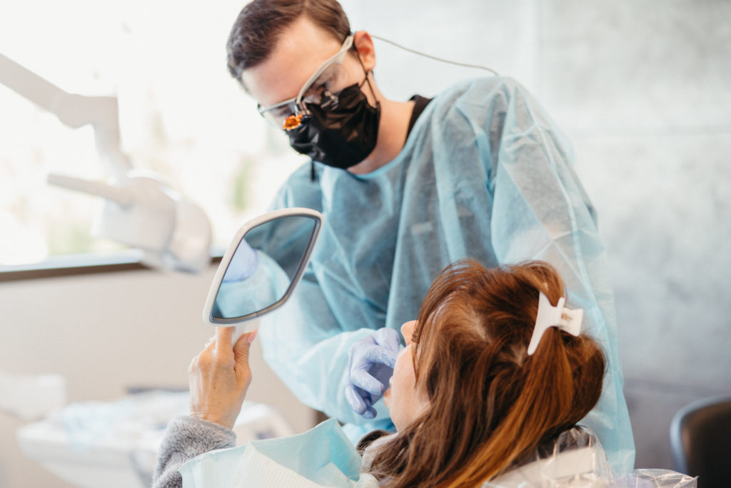 Dentist in protective gear showing a female patient her teeth in a mirror during a dental examination.
