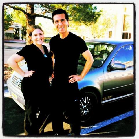 A man and a woman in black outfits standing smiling next to a silver car in a sunlit parking lot.