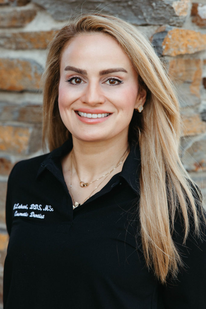 Portrait of a smiling woman with long blonde hair, wearing a black shirt with embroidered text, standing against a stone wall background.