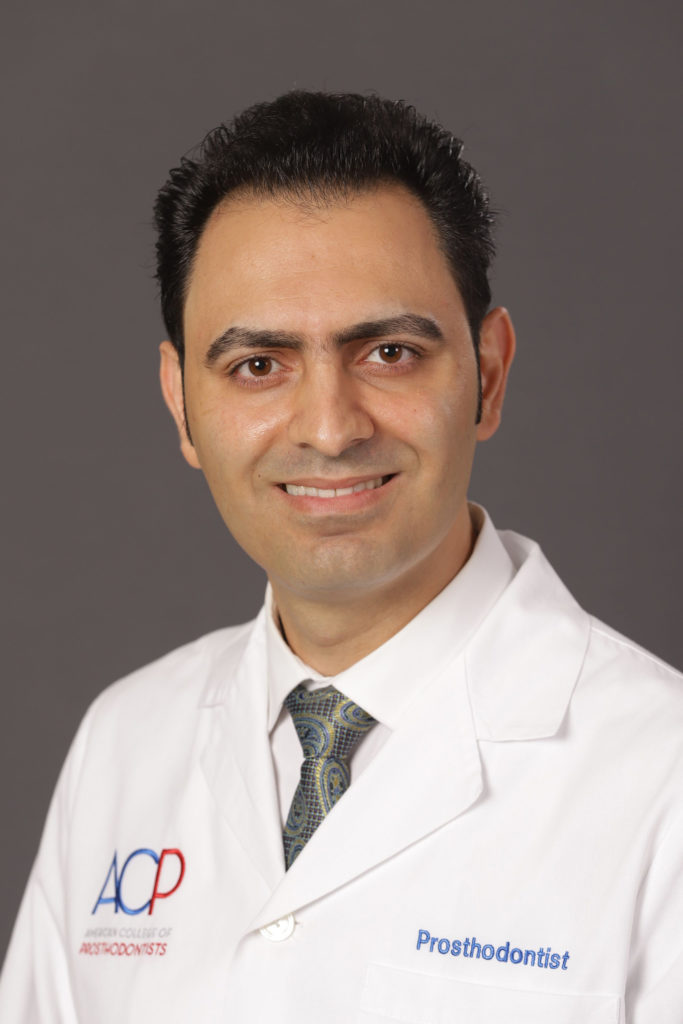Portrait of a smiling male prosthodontist wearing a white lab coat with an "acp - american college of prosthodontists" logo and a tie, against a gray background.