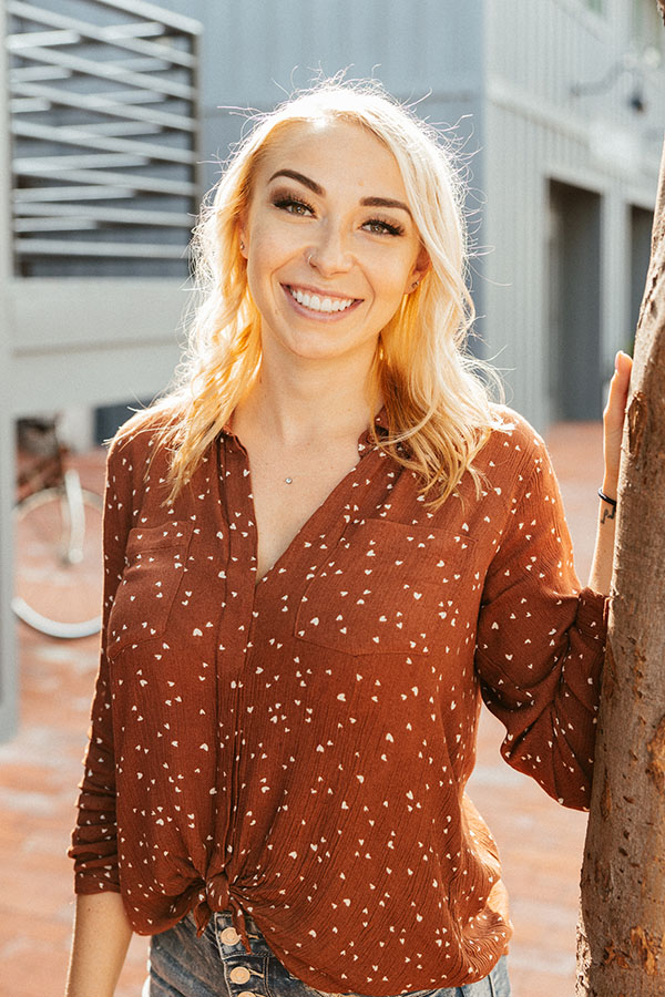 A smiling woman with blonde hair, enhanced by cosmetic dentistry, wearing a brown blouse, standing outdoors next to a tree.