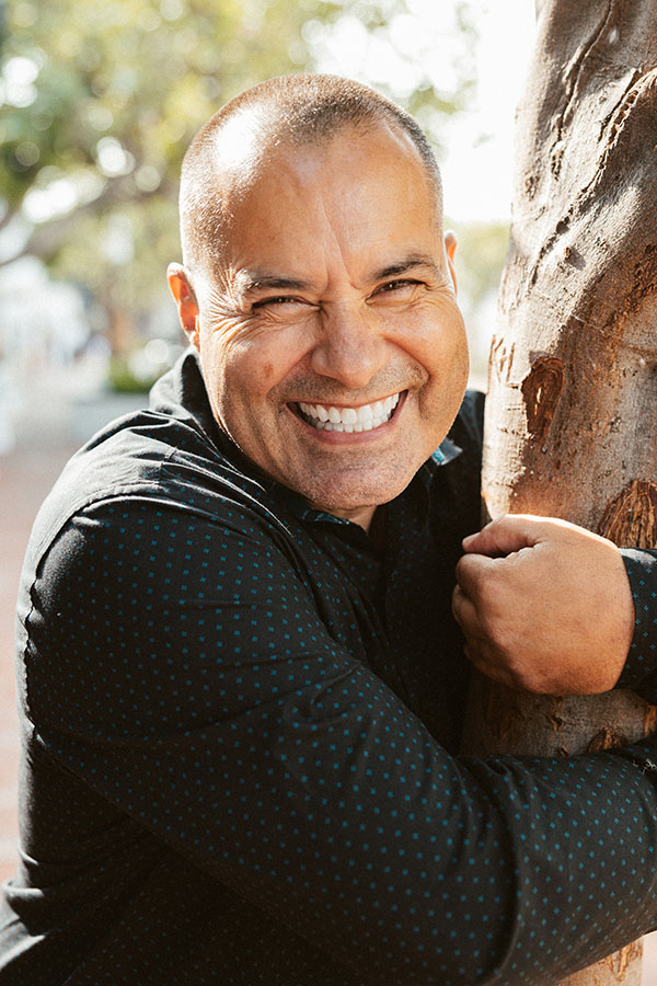 A smiling man with a cosmetic dentistry-enhanced smile embracing a tree on a sunny day, wearing a black shirt with blue dots.