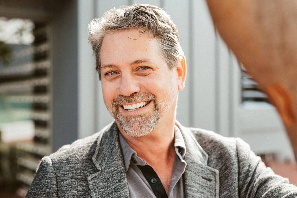 A smiling middle-aged man, exhibiting his cosmetic dentistry, with salt-and-pepper hair and a beard, wearing a gray blazer, outdoors in a sunny setting.