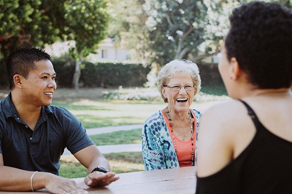 A joyful elderly woman laughs while conversing with a young man and a woman at an outdoor table, in a sunny park setting.