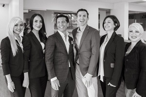 Six professionals smiling for a group photo in formal business attire, standing indoors in black and white.
