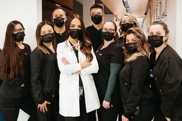 A group of nine diverse healthcare professionals, specializing in whole mouth dental implants, wearing black masks and dressed in black and white, standing confidently in a clinic hallway.
